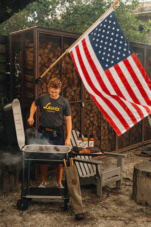 Grilling sausages under the USA flag in a GRILL KILT Grilling apron and Lewis Barbecue shirt